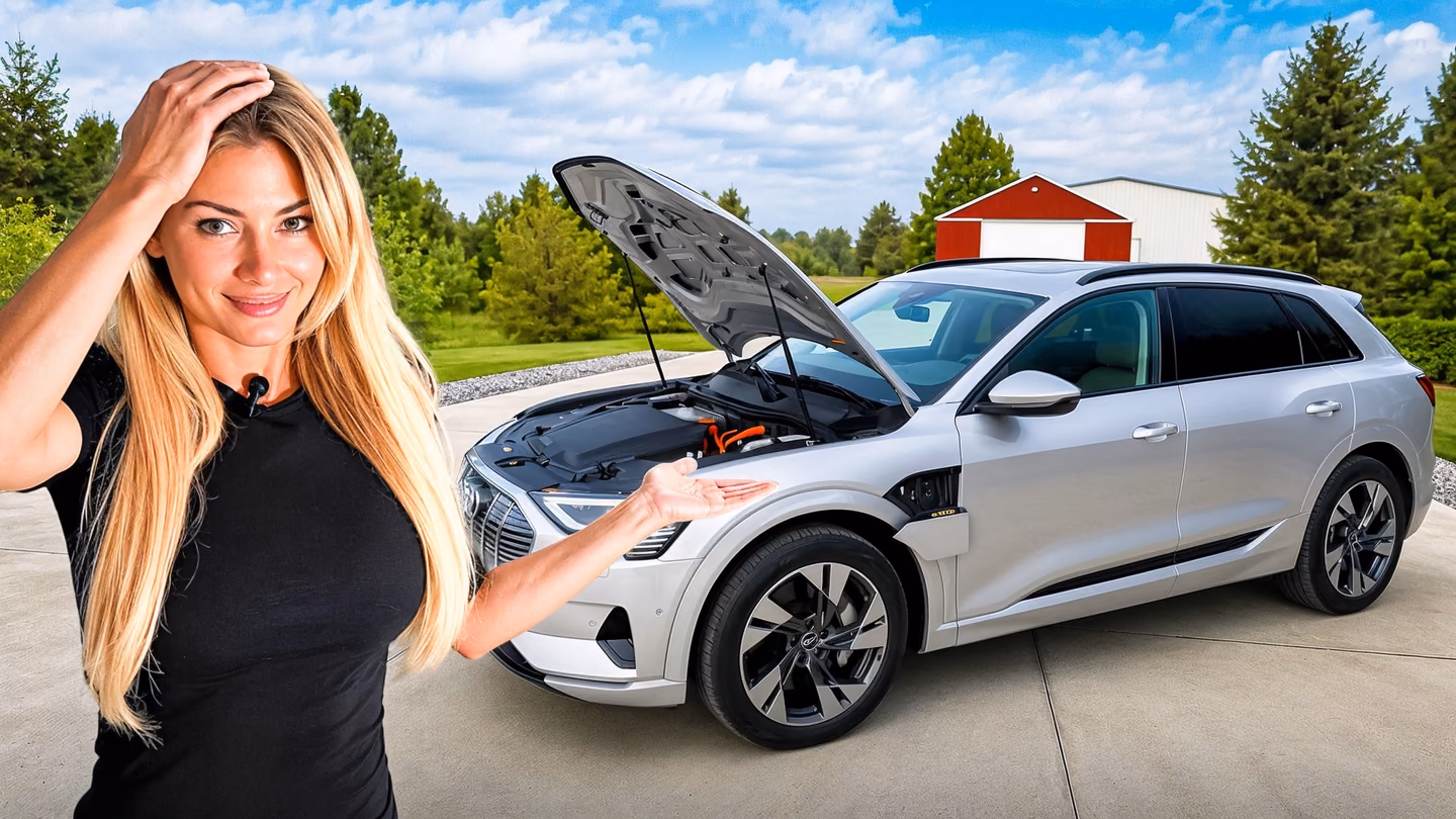 A woman gestures toward a silver Audi with its hood open in a bright driveway.